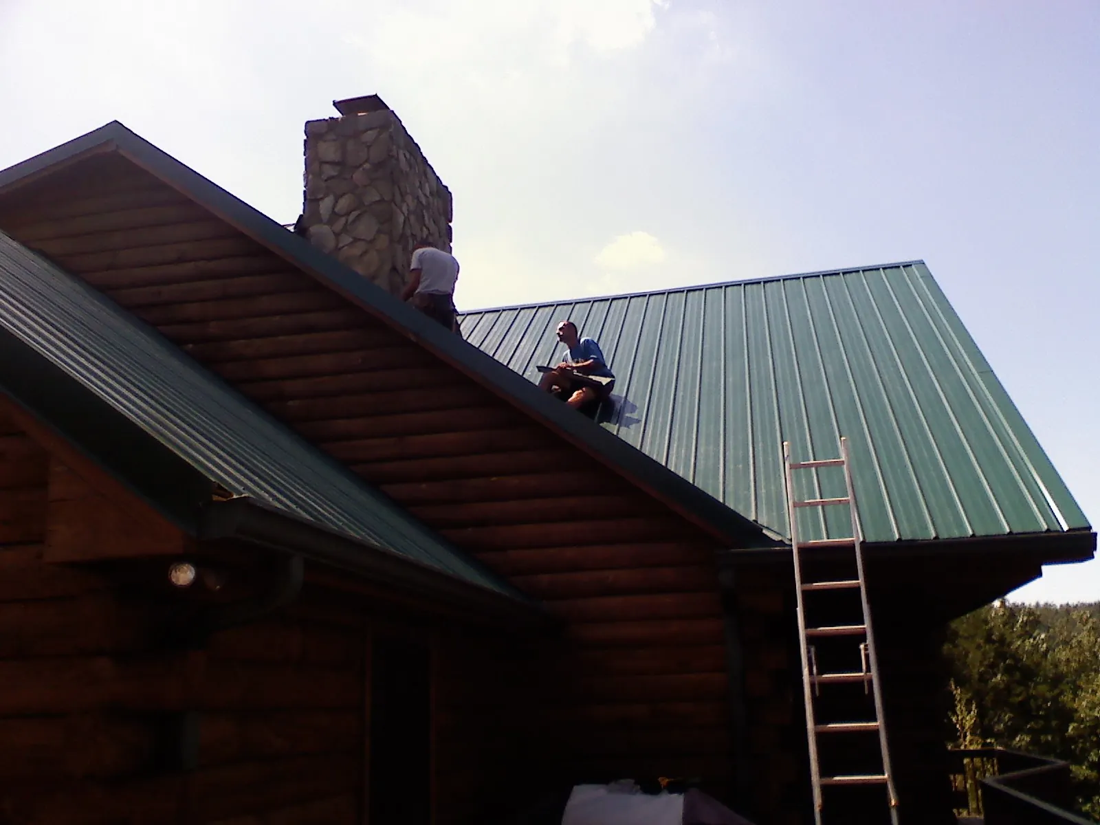 Roofers installing green metal panels beside a stone chimney on a log home in Virginia City